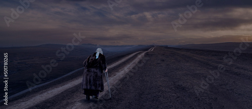A lonely elderly woman with a cane on a trampled road in the mountains. 