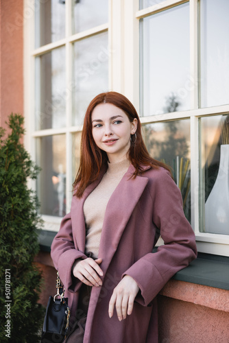 redhead young woman in coat standing near windows outside.