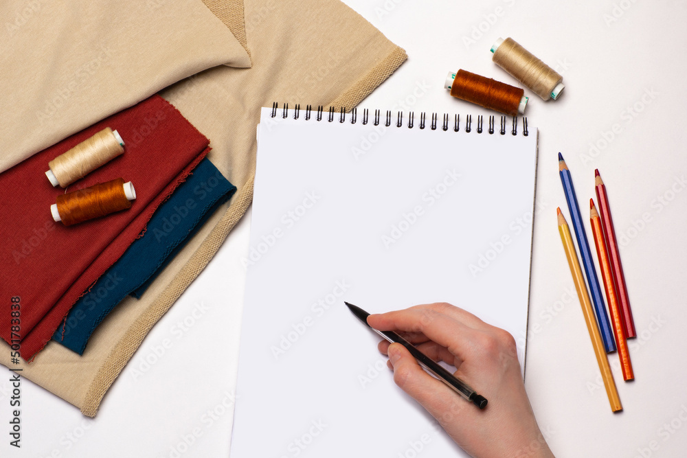 Mock up of white notebook and hand with pen on it. Sewing threads, fabric and pencils on white background. Mock up of white notebook and hand with pen on it. Sewing threads, fabric and pencils on white background.