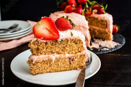 Slice of Fresh Strawberry Cake on a White Plate: A slice of cake on a small plate with the rest of the cake in the background