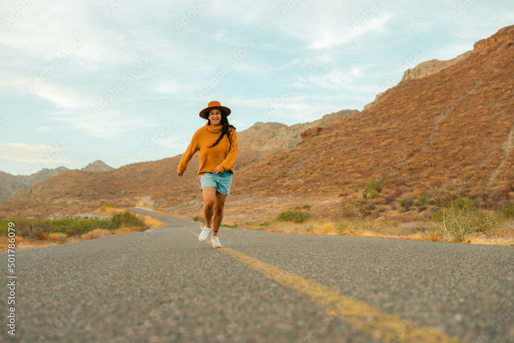 Cheerful young latin woman running, feeling free in the middle of a lonely road in the mountains of Baja California Sur. copy space.
