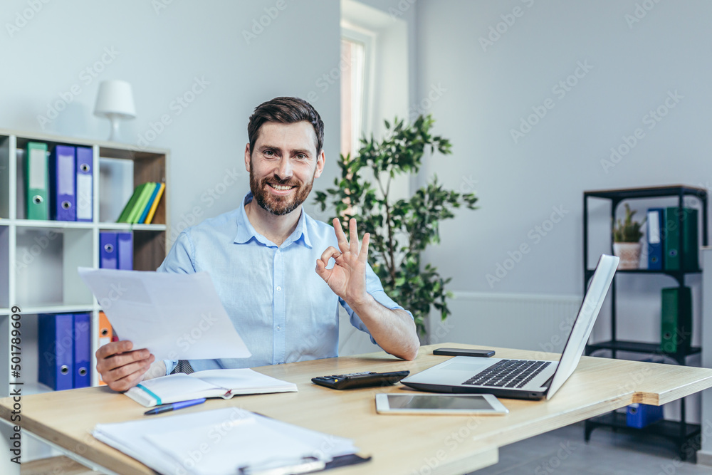 Successful businessman looks at the camera and smiles, performs reconciliation of business reports, shows the camera gesture ok Successful businessman looks at the camera and smiles, performs reconciliation of business reports, shows the camera gesture ok