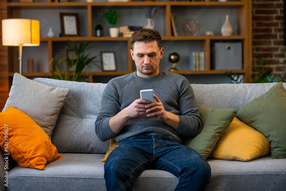 Portrait of an attractive serious young man wearing casual clothes sitting on a couch at the living room, using mobile phone