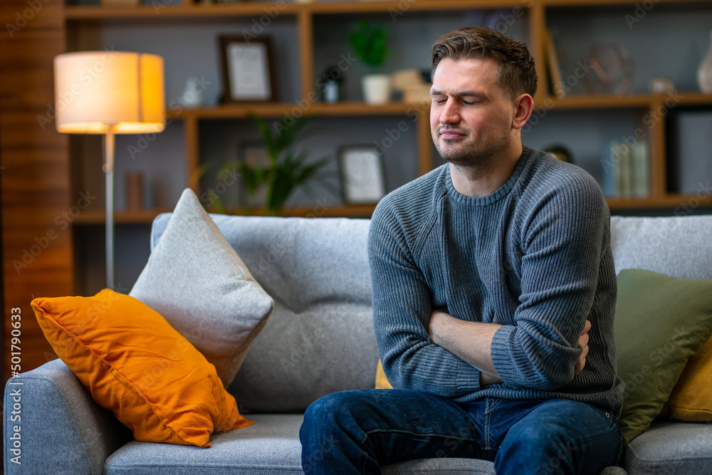 A young man suffering from abdominal pain holding his stomach, sitting on the sofa. A young man suffering from abdominal pain holding his stomach, sitting on the sofa.