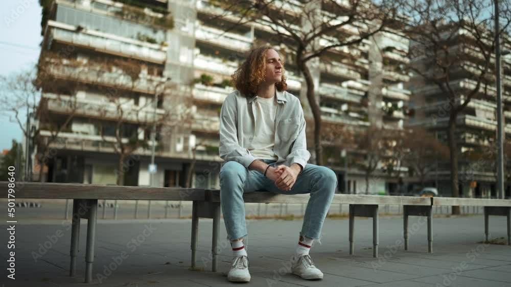 Concentrated curly-haired man looking at the view while sitting on the bench outdoors Concentrated curly-haired man looking at the view while sitting on the bench outdoors