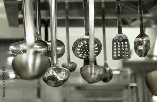 A view of several metal ladles and large mixing spoons hanging in a restaurant kitchen setting
