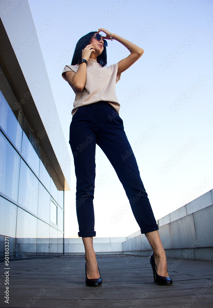 teen businesswoman. young stylish girl with long legs stands casual with mobile phone near ear and looks apart on glass business centre background with blue sky. lifestyle business concept, free space