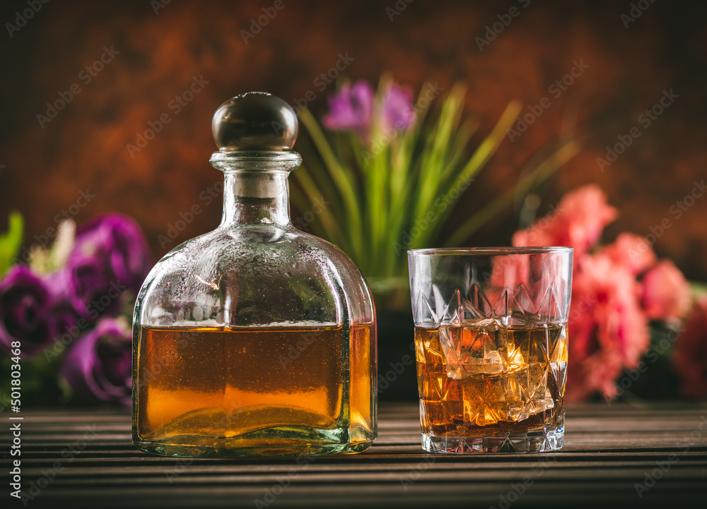 Glass of whiskey with ice and bottle with liquor on a wooden table with some color flowers. Alcoholic beverage ready to drink. Glass of whiskey with ice and bottle with liquor on a wooden table with some color flowers. Alcoholic beverage ready to drink.