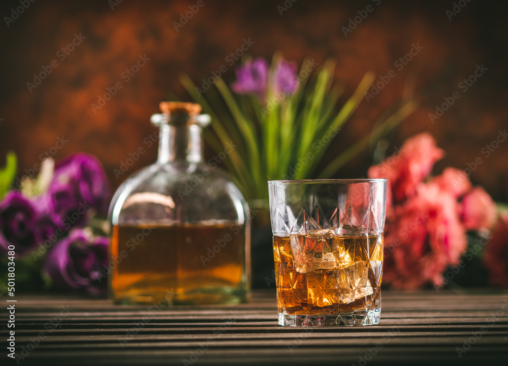 Glass of whiskey with ice and bottle with liquor on a wooden table with some color flowers. Alcoholic beverage ready to drink. Glass of whiskey with ice and bottle with liquor on a wooden table with some color flowers. Alcoholic beverage ready to drink.