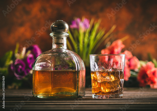 Glass of whiskey with ice and bottle with liquor on a wooden table with some color flowers. Alcoholic beverage ready to drink.