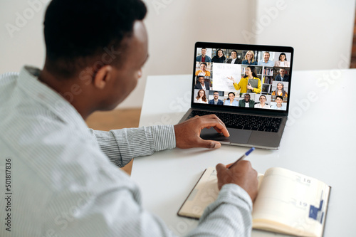 Online learning. African American smart guy, student or freelancer, sit at a table at home in the kitchen, has an online webinar, lecture, takes notes, on the screen group of multiracial people