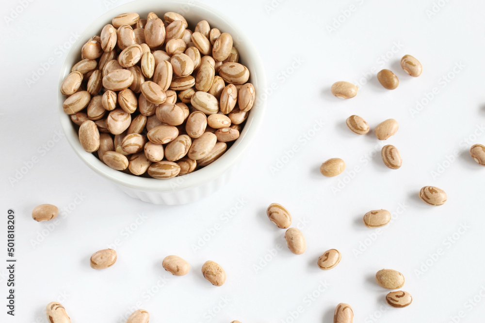 Carioca beans in a white bowl with pinto grains scattered on white background. Carioca beans in a white bowl with pinto grains scattered on white background.
