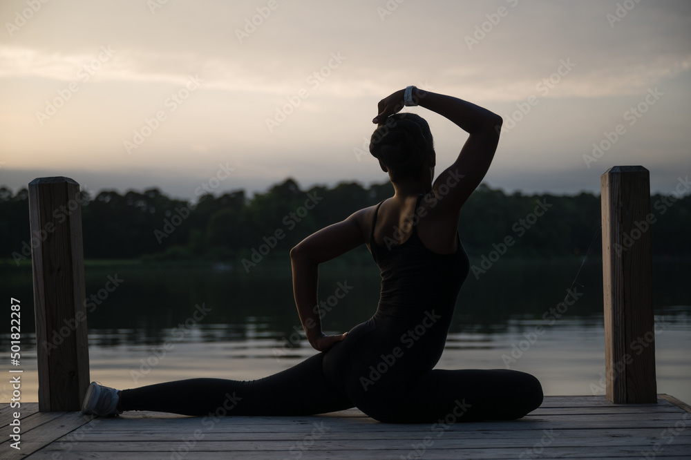 a slim young African American woman stretches at sunset on a pier in front of a lake one summer