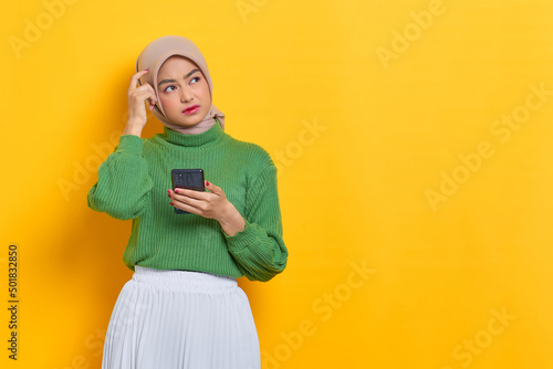 Pensive beautiful Asian woman in green sweater holding mobile phone looks seriously thinking about something isolated over yellow background