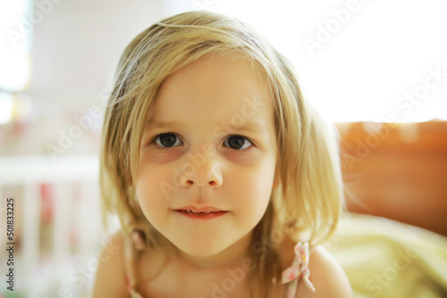 A small child with blond hair lies on the bed. Baby girl is playing on the couch.