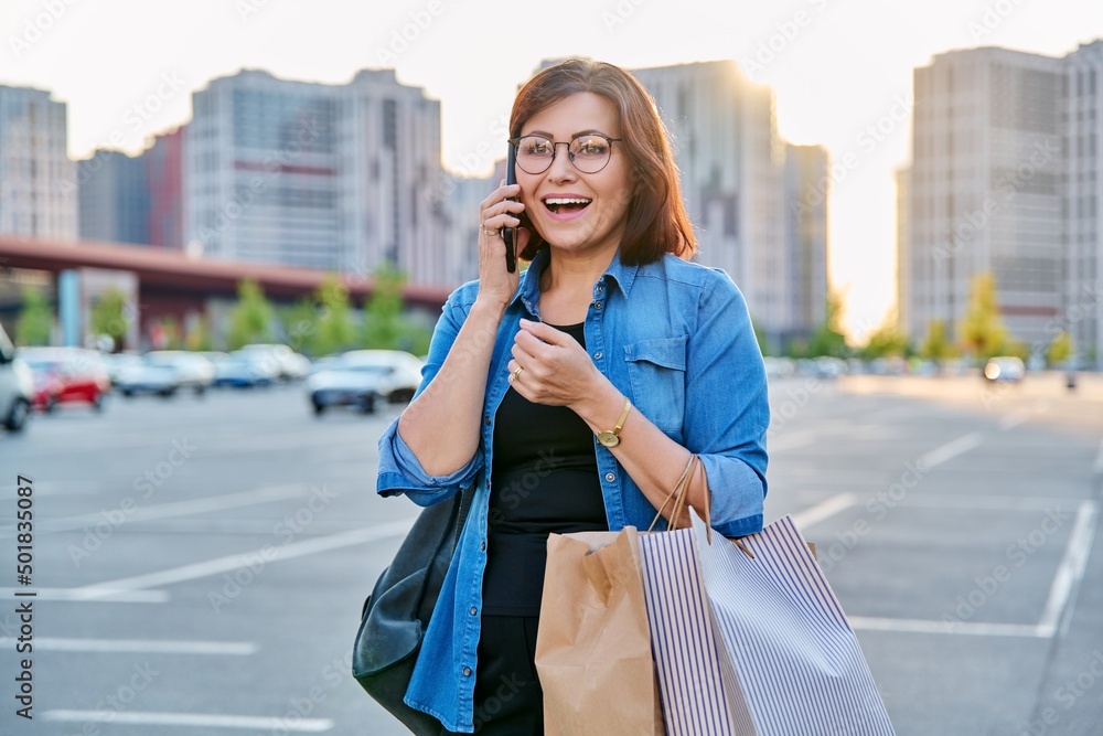 Fototapeta premium Outdoor portrait of a beautiful 40s woman talking on phone with shopping bags