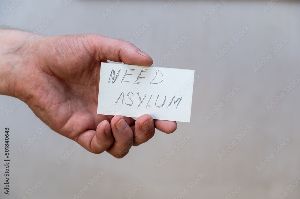 Need asylum. Words written in jagged letters. A man's hand holds a white paper rectangle with text against a gray background. Need asylum. Words written in jagged letters. A man's hand holds a white paper rectangle with text against a gray background.