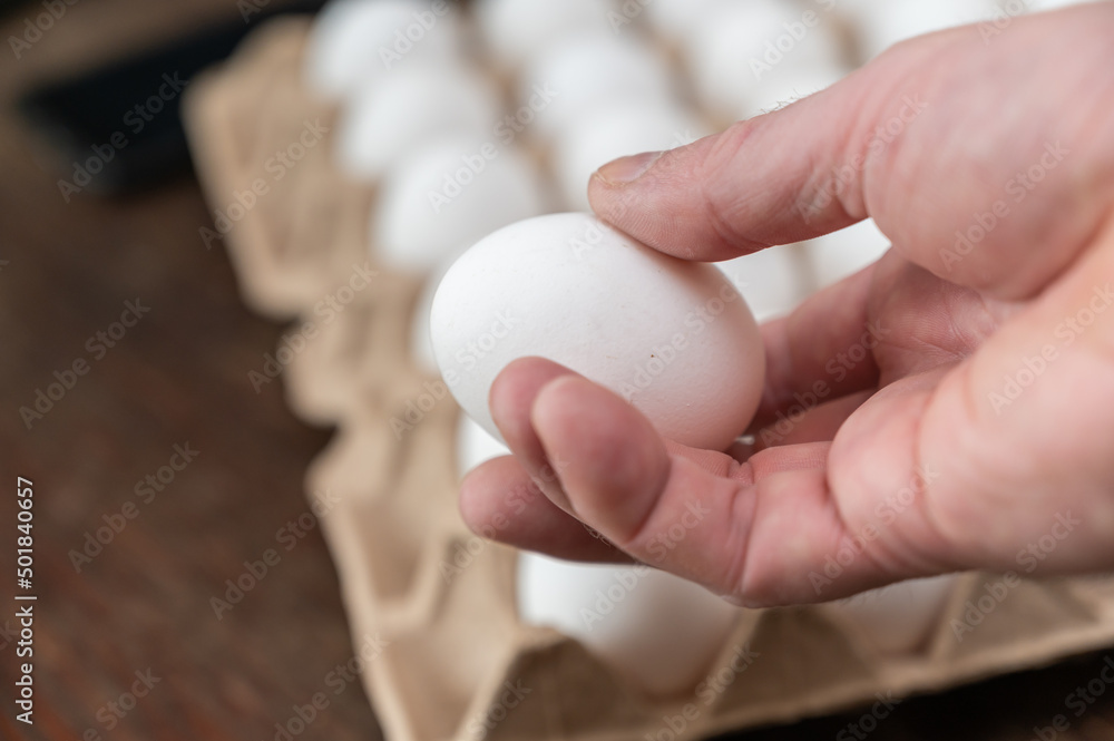 A man is holding a chicken egg in front of an egg tray. Adult male holding a white raw egg. Open recycled tray. Selective Focus. A man is holding a chicken egg in front of an egg tray. Adult male holding a white raw egg. Open recycled tray. Selective Focus.