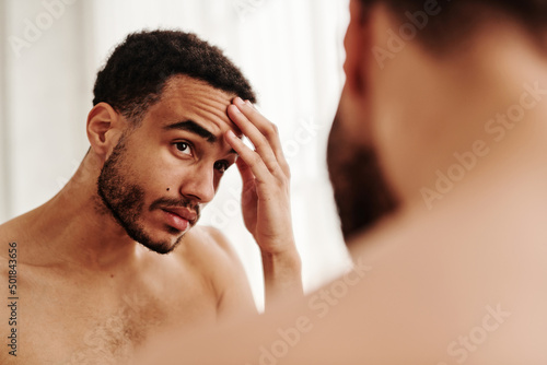 Young handsome man examining wrinkles on his face while looking at mirror photo