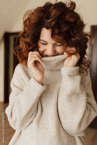 Charming cute lady with curls wearing knitted pullover is laughing and looking down with closed eyes. Indoor portrait of romantic pretty lady