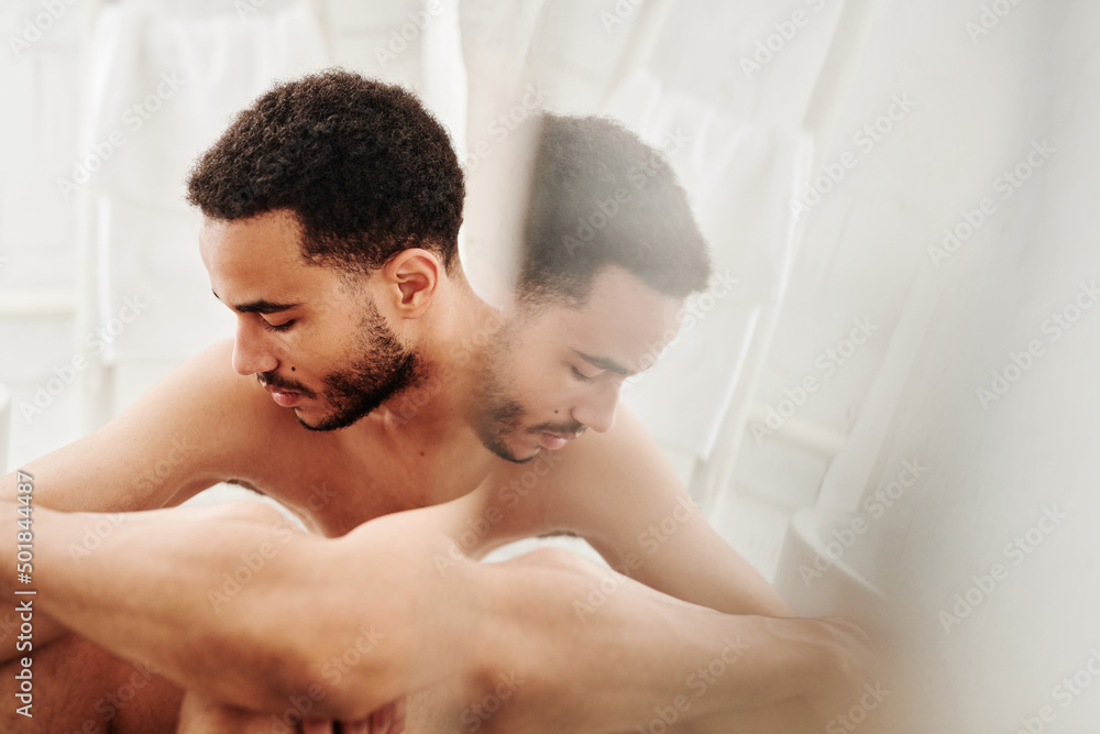 Fototapeta premium Young shirtless man sitting in front of mirror with sad expression and thinking about something