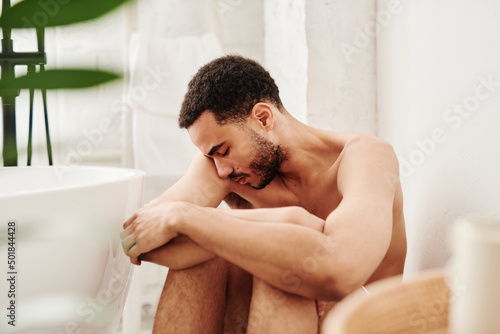 Young shirtless man sitting on floor in bathroom alone and having depression