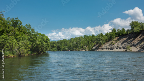 A calm blue river flows under an azure sky. The steep banks are covered with green vegetation. An inflatable rafting boat with people is visible in the riverbed. A sunny summer day. Kamchatka