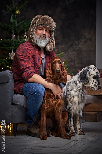 Portrait of grandfather with hat sitting on chair with his canine pets indoors room decorated with christmas ornaments.