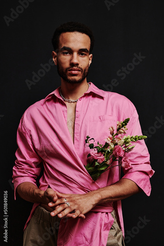 Portrait of young mixed race man in pink shirt with bouquet of flowers inside looking at camera against black background