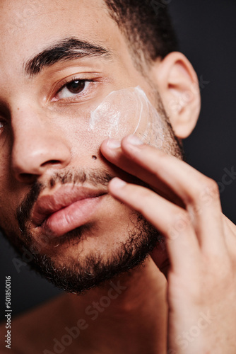 Close-up of bearded young man applying face cream on his face and looking at camera
