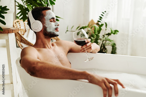 Young man enjoying relaxed time in bathroom, he listening to music in wireless headphones and enjoying the glass of red wine photo