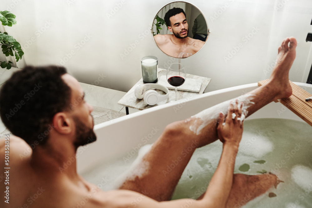 Rear view of young man applying shave foam on his leg while taking a bath in bathroom Rear view of young man applying shave foam on his leg while taking a bath in bathroom