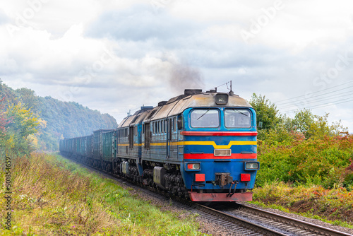 A powerful blue diesel locomotive pulls a long freight train loaded with coal along the railway tracks. Autumn photo. International Freight rail transport