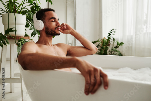 Fotografiet Young handsome man in wireless headphones relaxing in bath with foam and enjoyin