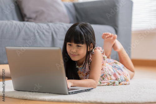 Photography Cute little asian girl uses laptop while prone at the sofa in the living room
