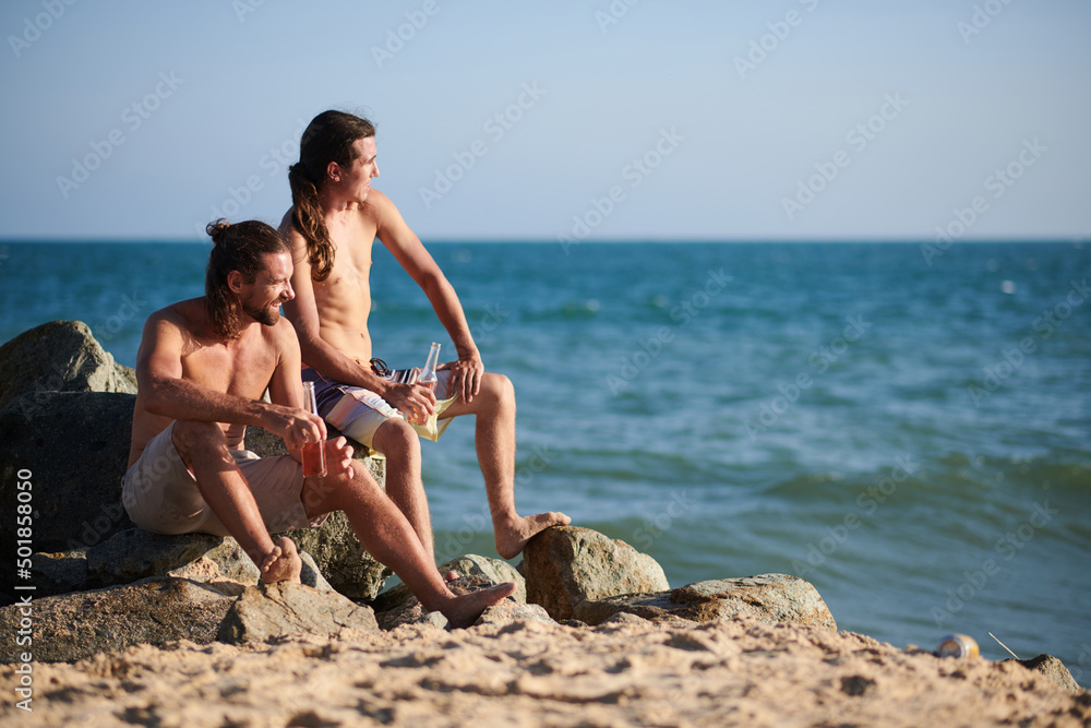 Men sitting on rock on beach, drinking refreshing beverages and looking at waves Men sitting on rock on beach, drinking refreshing beverages and looking at waves