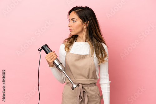 Young woman using hand blender over isolated pink background looking to the side