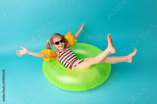 Happy little child girl in a swimwear suit lying on a bright inflatable circle. Blue background. Top view. Summer and vacation concept