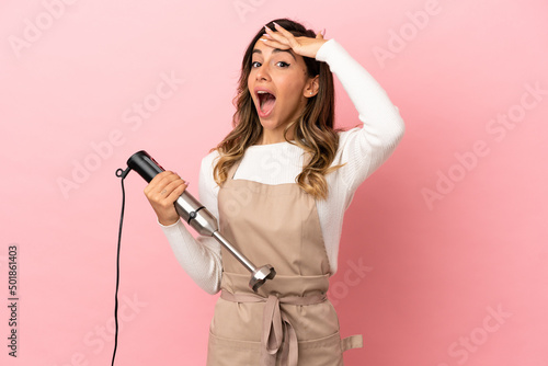 Young woman using hand blender over isolated pink background doing surprise gesture while looking to the side