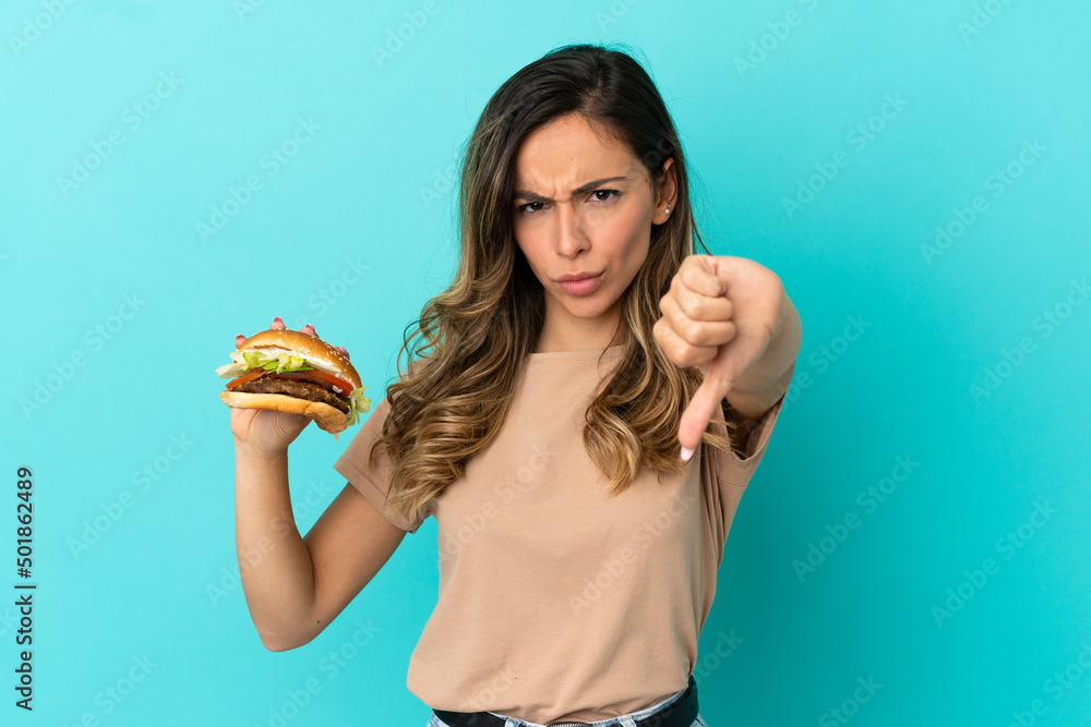 Young woman holding a burger over isolated background showing thumb down with negative expression Young woman holding a burger over isolated background showing thumb down with negative expression