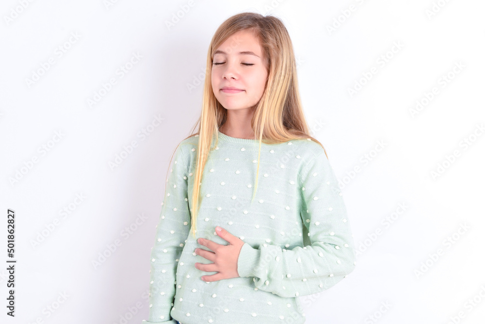 little caucasian kid girl wearing fashion sweater over blue background touches tummy, smiles gently, eating and satisfaction concept. little caucasian kid girl wearing fashion sweater over blue background touches tummy, smiles gently, eating and satisfaction concept.