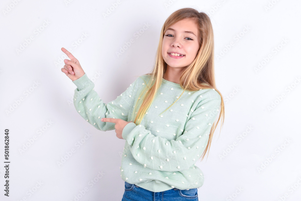 little caucasian kid girl wearing fashion sweater over blue background indicating finger empty space showing best low prices, looking at the camera little caucasian kid girl wearing fashion sweater over blue background indicating finger empty space showing best low prices, looking at the camera