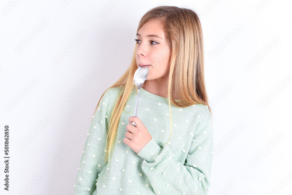 Very hungry little caucasian kid girl wearing fashion sweater over blue background holding spoon into mouth dream of tasty meal Very hungry little caucasian kid girl wearing fashion sweater over blue background holding spoon into mouth dream of tasty meal