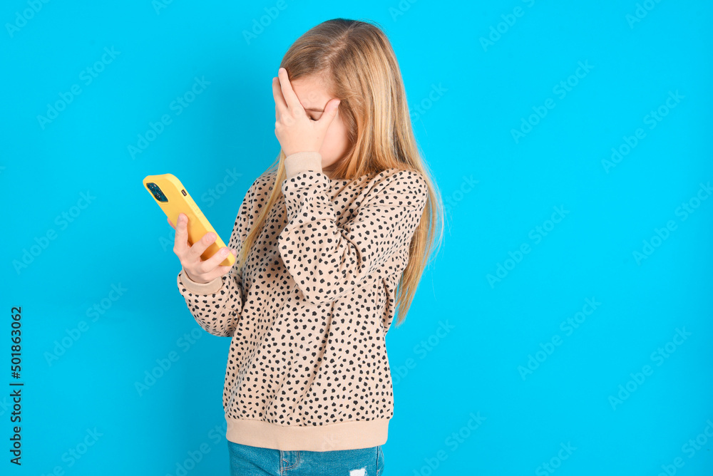 little caucasian kid girl wearing animal print sweater over blue background looking at smart phone feeling sad holding hand on face. little caucasian kid girl wearing animal print sweater over blue background looking at smart phone feeling sad holding hand on face.