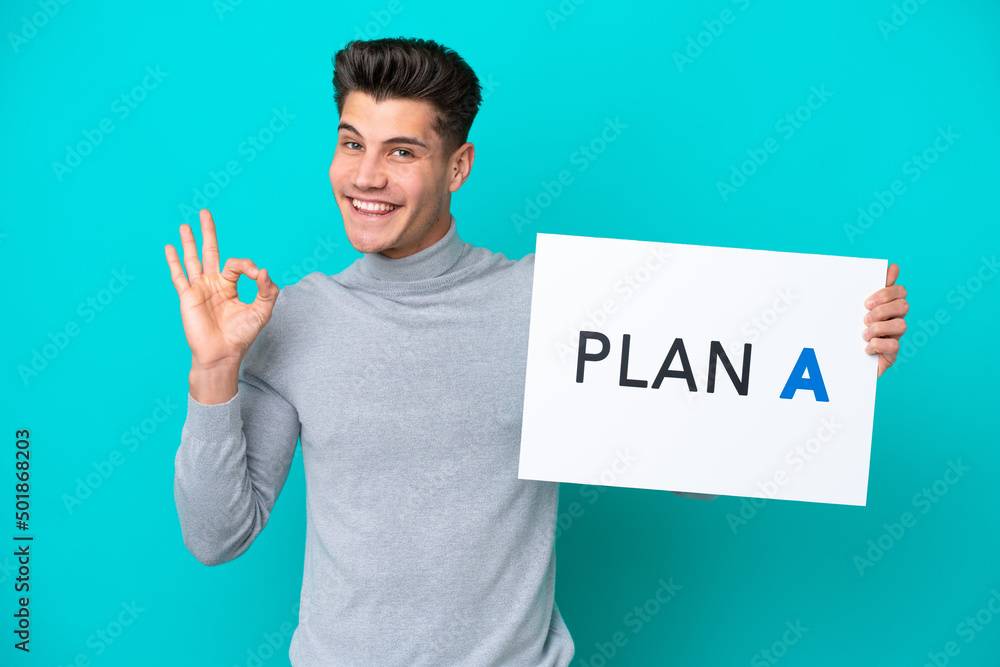 Young handsome caucasian man isolated on blue bakcground holding a placard with the message PLAN A with ok sign Young handsome caucasian man isolated on blue bakcground holding a placard with the message PLAN A with ok sign