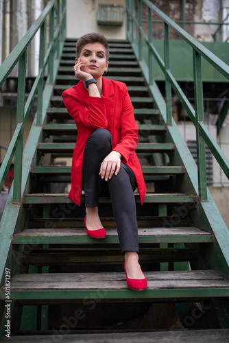 A stylish model girl with short brown hair in a red jacket and pumps, a white T-shirt, posing for a photographer sitting on a wooden staircase with wooden railings and propping her face with her hand