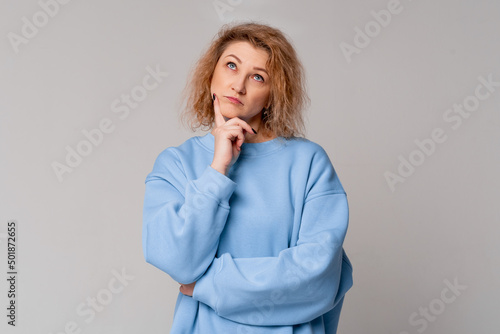 Thoughtful middle age woman with curly blonde hair remembering something, standing with doubtful expression in trendy blue sweatshirt over light grey background. Let me think. Doubt concept