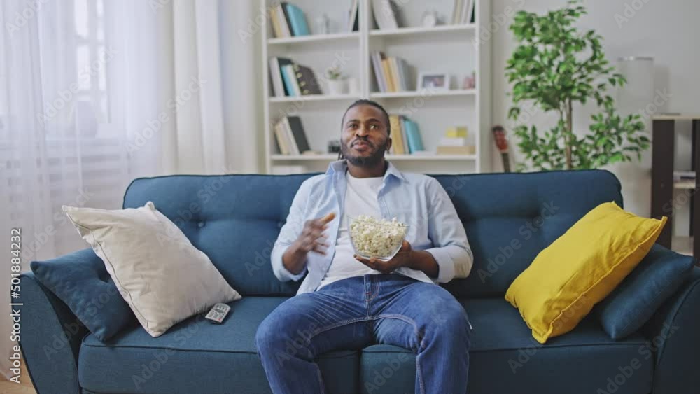 African American man sitting on sofa, watching movie on TV and eating popcorn African American man sitting on sofa, watching movie on TV and eating popcorn
