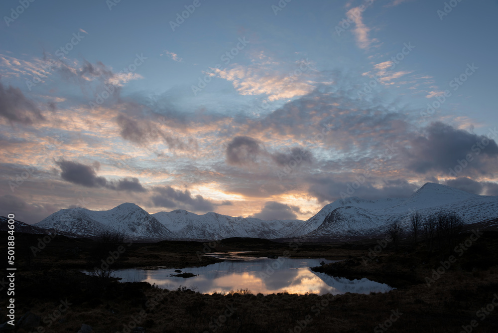 Epic Winter sunset landscape image across Loch Ba in Scottish Highlands towards snow covered mountain range in distance