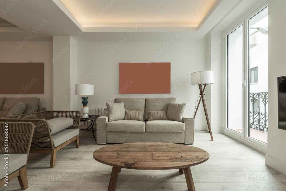 Living room with oval unvarnished wooden table on wooden floors, balcony with metal railings and gray fabric and rattan sofas Living room with oval unvarnished wooden table on wooden floors, balcony with metal railings and gray fabric and rattan sofas
